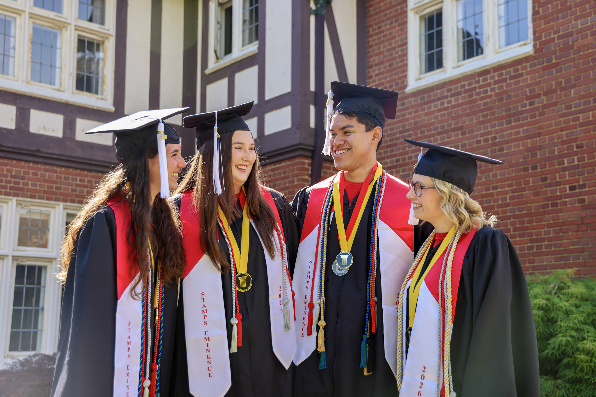 Four graduates are standing together outside Kuhn House smiling wearing caps and gowns with red and white stoles and various cords.