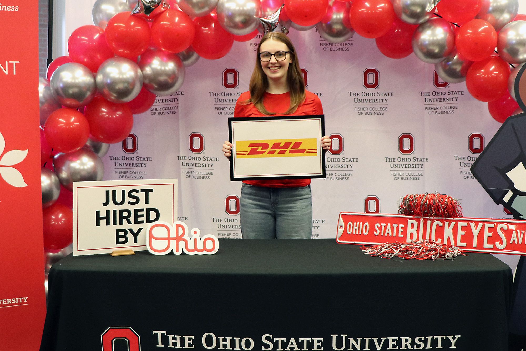 A person stands smiling behind a table at The Ohio State University, decorated with red and silver balloons. They hold a sign with the DHL logo.