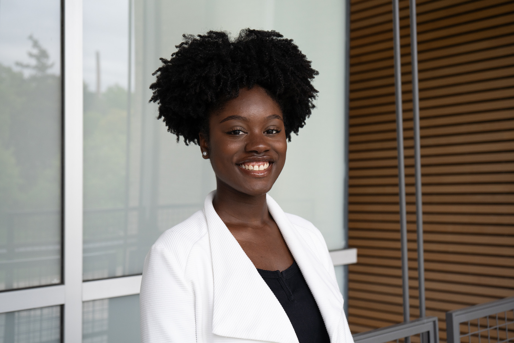 Chelsea smiles in white jacket with black shirt with a window and wooden wall behind her