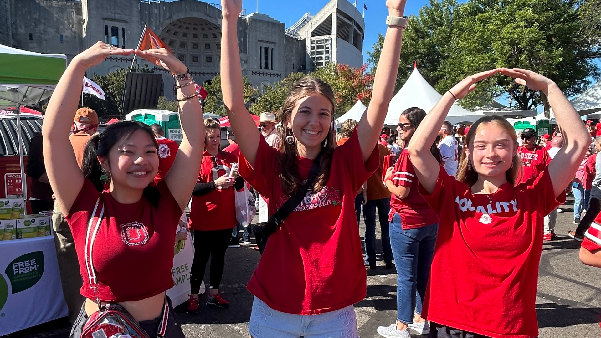 Three people forming the letters "O-H-I-O" with their arms at Ohio Stadium in the background with others in the area