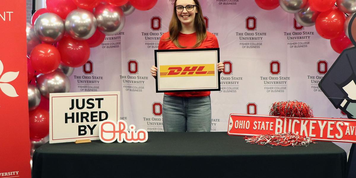 A person stands smiling behind a table at The Ohio State University, decorated with red and silver balloons. They hold a sign with the DHL logo.