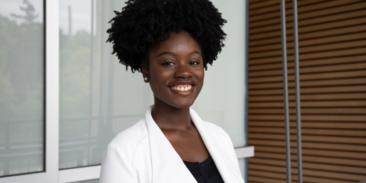 Chelsea smiles in white jacket with black shirt with a window and wooden wall behind her