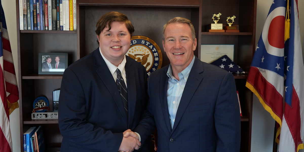 Two people in suits are shaking hands in an office with a bookshelf behind them. 