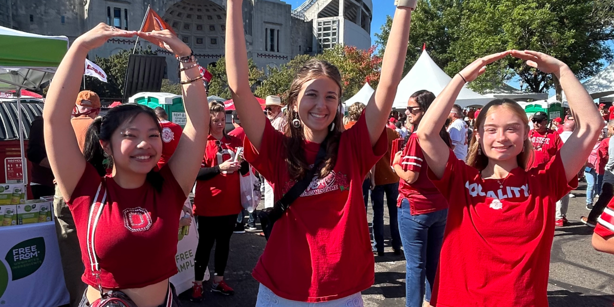Three people forming the letters "O-H-I-O" with their arms at Ohio Stadium in the background with others in the area