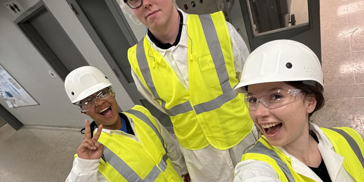 Three people wearing white hard hats, safety goggles, and yellow reflective vests stand in a hallway with gray doors and a bulletin board.