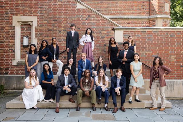  Group photo of twenty people dressed in formal and semi-formal attire, posing in front of a brick building.