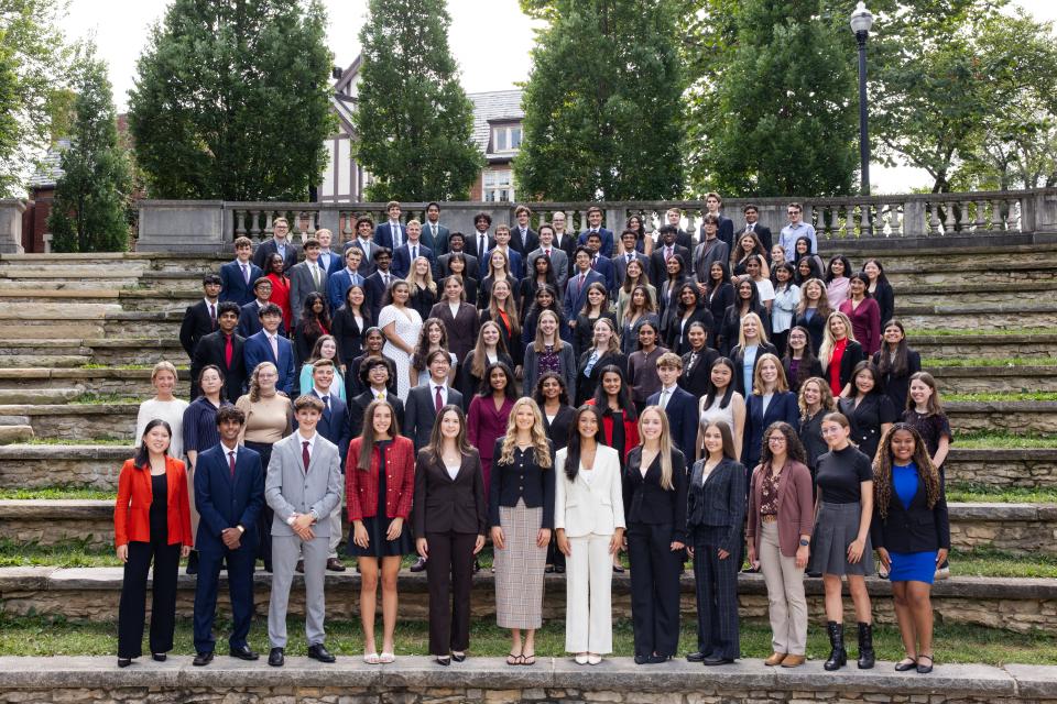 A large group of people dressed in formal attire are gathered on outdoor stone steps. The setting is surrounded by greenery and trees.