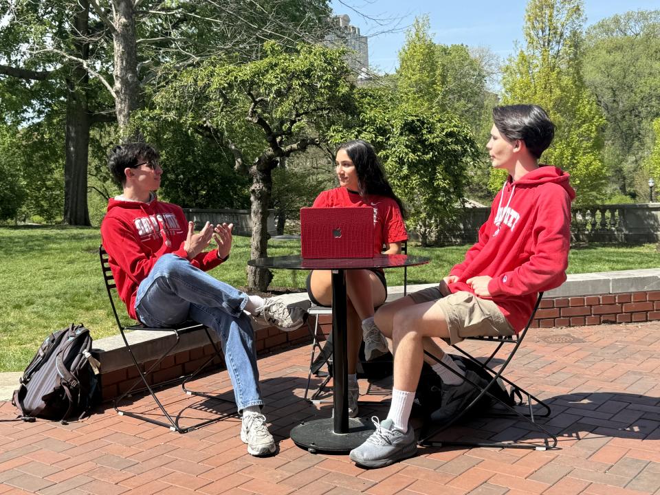 Three people are seated outdoors at a small table with a laptop open with a backpack on the ground. The setting is a sunny, leafy area with brick paving.