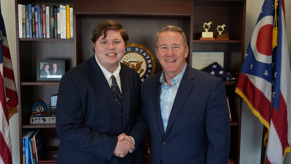 Two people in suits are shaking hands in an office with a bookshelf behind them.