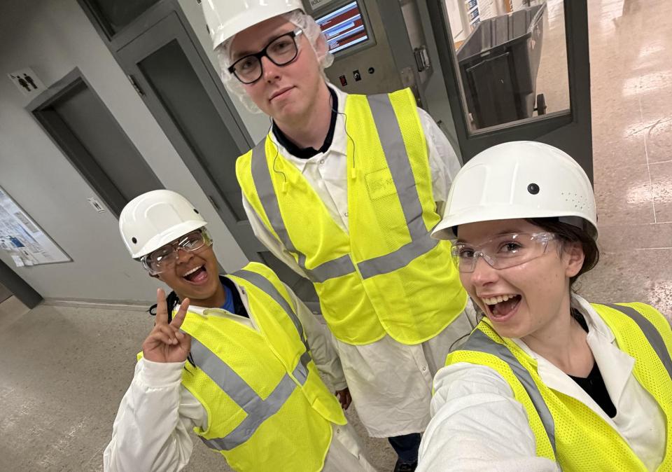 Three people wearing white hard hats, safety goggles, and yellow reflective vests stand in a hallway with gray doors and a bulletin board.