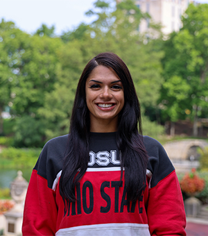 A person smiling in an outdoor setting, wearing an Ohio State sweatshirt with green trees and a building in the background.