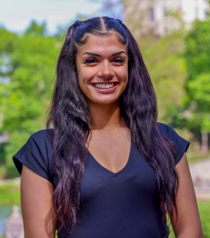 A person is smiling, wearing a black top, standing outdoors with a blurred background of greenery and buildings.